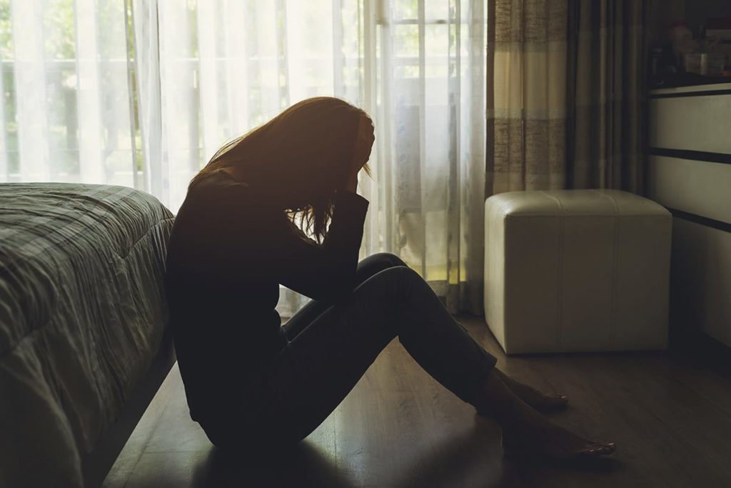 a woman sits on the floor by the foot of her bed with her head in her hands sick from meth abuse