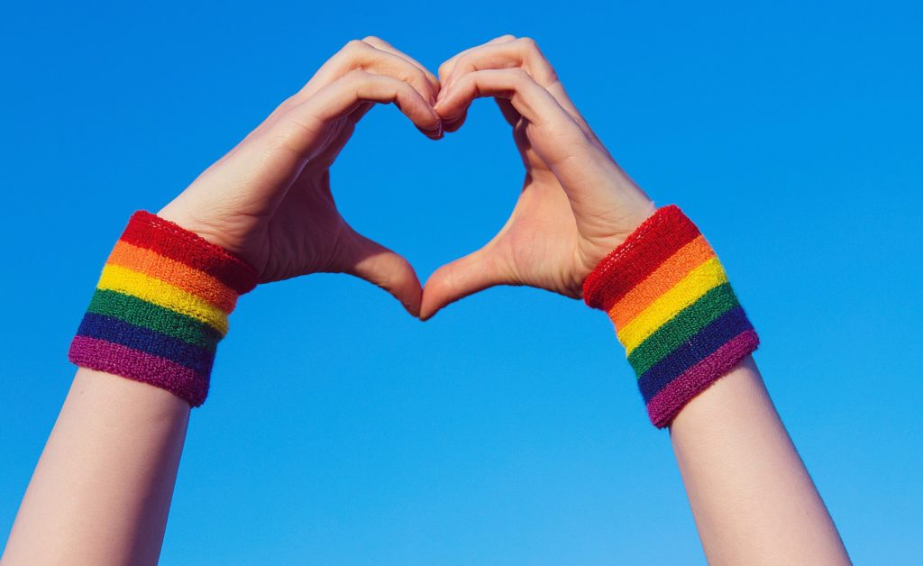 Hands reaching up making a heart with the fingers. Each wrist has a rainbow sweatband on it.