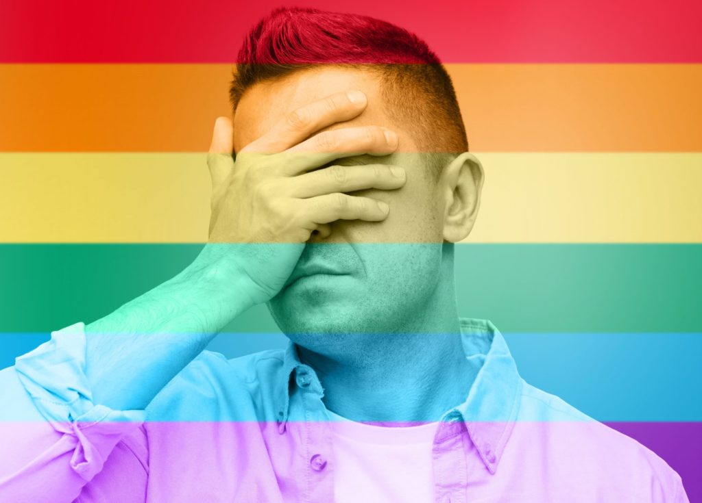 A man covers his eyes with the palm of his hand. There is a rainbow filter over the picture.