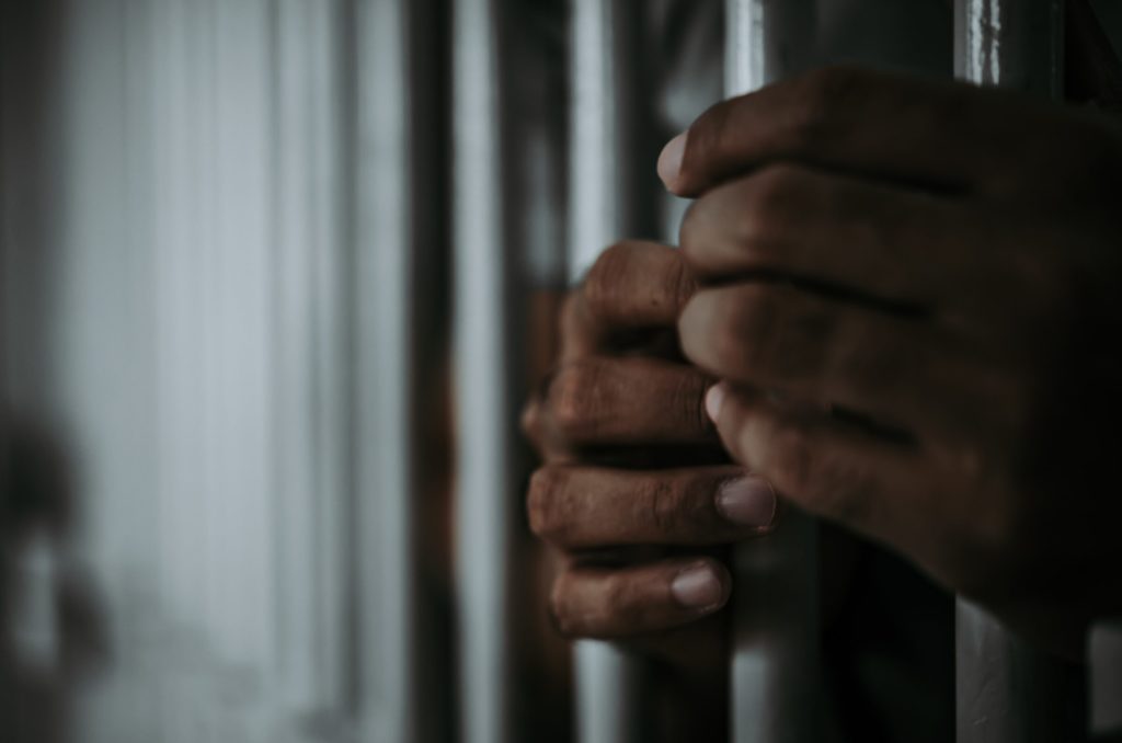 A close up of an African American's hands holding bars inside of a jail cell.  