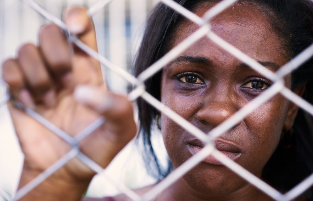 A close up of an American American woman behind a chain-link fence. One hand is holding the fence. 