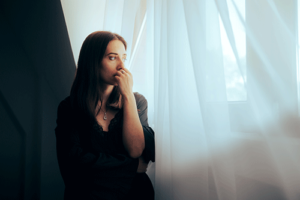 Young woman standing nervously by a curtained window looking pensive and uncertain as she contemplates a prescription drug detox program
